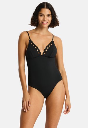 Donna con capelli corti e ricci sorridente, che indossa un costume intero nero con bordi smerlati e dettagli di ritagli circolari sullo scollo.