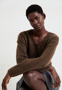 Cardigan marrone peloso con scollo a V e chiusura con bottoni, abbinato a una gonna nera. Modella posizionato seduta, braccia incrociate, che mette in mostra la texture del tessuto.