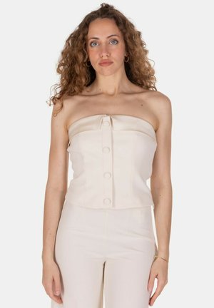 Donna con capelli ricci che indossa un top beige senza spalline con bottoni e pantaloni a vita alta abbinati, in piedi davanti a uno sfondo bianco semplice.