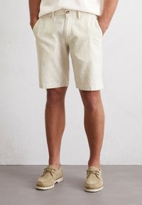 Beige cotton shorts with a vertical stripe pattern, relaxed fit, side pockets, and a single button closure. Worn with light-colored shoes.