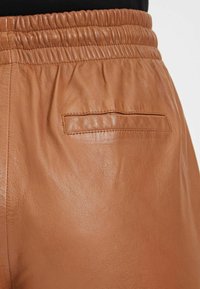 Bruine leren broek met een gestructureerde afwerking, elastische tailleband en een enkele achterzak zonder zichtbare hardware.