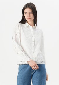 White cotton blouse with intricate eyelet pattern, long sleeves, button front, and scalloped cuffs, paired with denim jeans.
