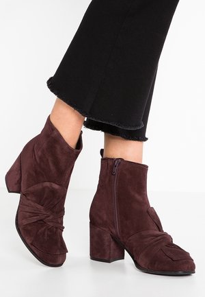 Brązowe zamszowe botki na słupku z bocznymi zamkami, noszone z czarnymi, skróconymi spodniami z postrzępionymi zakończeniami.