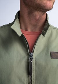 Zelená nylonová bunda se stojáčkem, plným zipem a koženou nášivkou. Nosená přes korálovou košili s texturovaným povrchem.