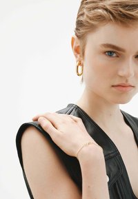 Top sin mangas de cuero negro, con ligero brillo y costuras visibles. Pendiente de aro dorado, pulsera delgada de oro en el brazo, tono de piel claro.