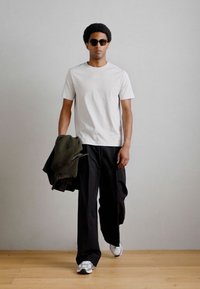 Biela tričko, čierne široké nohavice, drží zelenú bundu a nosí slnečné okuliare. Tenisky sú metalicky strieborné s hladkou textúrou.