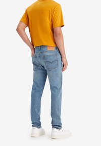 Jeans in denim azzurro chiaro con vestibilità dritta, caratterizzati da tasche posteriori e un'etichetta in pelle. Abbinati a una maglietta a maniche corte giallo senape e sneaker bianche.