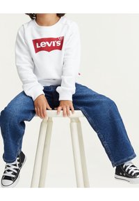 Vit sweatshirt med röd Levi's-logga, blå denimbyxor och svarta sneakers. Barn sitter på en pall, med händerna vilande på knäna.