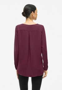 Blusa bordeaux con maniche lunghe, dettaglio della piega posteriore, orlo arrotondato e polsini con bottoni, realizzata in tessuto leggero. Vengono indossati pantaloni neri.