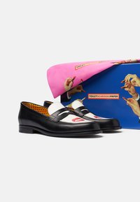 Crne i bijele kožne mokasinke s grafikom ruža za usne i usana na vrhovima, izložene ispred šarene kutije i ružičaste tkanine s ilustracijama ruke i ruža za usne.