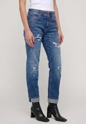 Person trägt blaue, ausgefranste Jeans mit hochgekrempelten Säumen, kombiniert mit schwarzen Stiefeletten mit Absatz und einem weißen ärmellosen Oberteil.