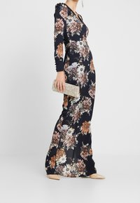 Woman wearing a long black floral dress with long sleeves, holding a sparkly clutch, and wearing beige pointed shoes.