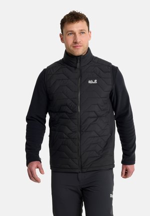 Mann trägt eine schwarze gesteppte Weste mit Reißverschluss über einem schwarzen Langarmshirt und steht vor einem schlichten hellen Hintergrund.
