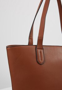Hnědá kožená taška s texturovaným povrchem, vybavená dvěma robustními rukojetěmi a černým akcentem podél horní hrany. Jednoduchý a moderní design.
