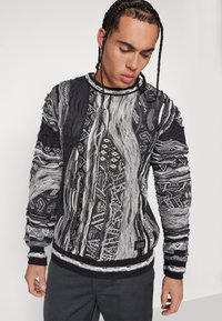 Gestrickter Pullover in Schwarz und Weiß mit einem Kabeldesign, strukturierten Mustern und kontrastierenden Rippbündchen und Saum. Logo-Tag unten links.