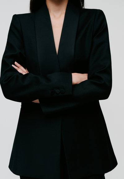Black blazer with a deep V-neck design, sleek texture, and tailored fit. Features three-button detailing on the cuffs.
