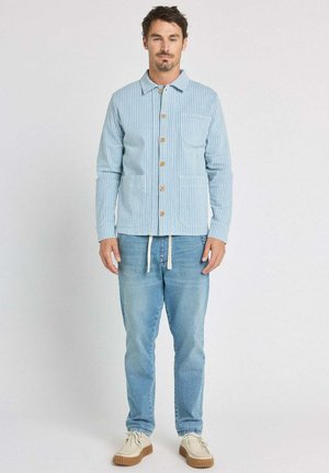 Homme debout face à l’avant portant une veste boutonnée rayée bleu clair, un jean bleu délavé avec des cordons blancs et des chaussures beiges.