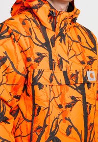 Giacca arancione con motivo di rametti neri, dotata di cappuccio e tasca frontale. Realizzata in un tessuto durevole e testurizzato.