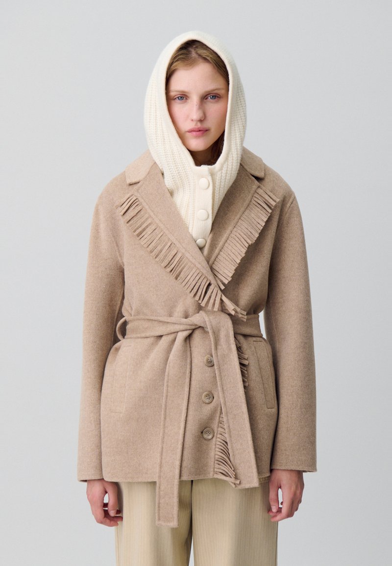 Femme portant un manteau ceinturé à franges beige sur un pull à capuche côtelé crème avec des boutons, se tenant contre un fond gris clair uni.