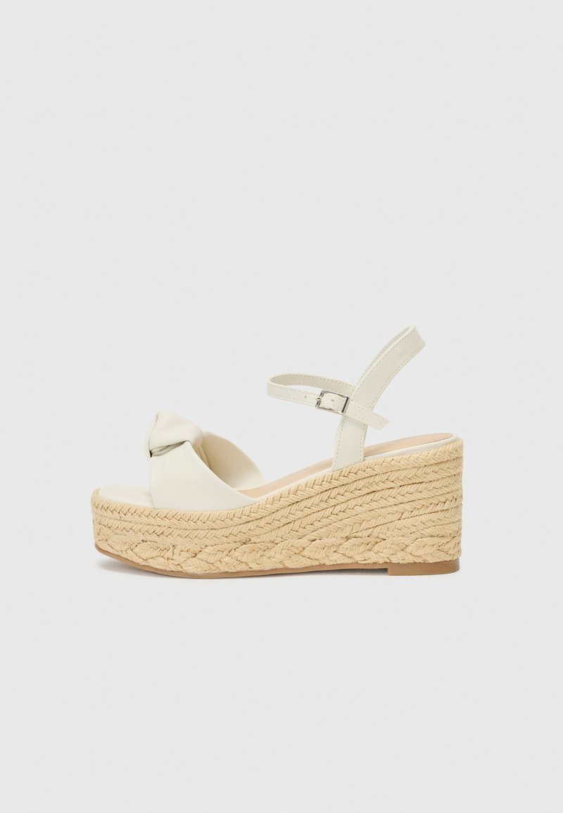 Cream wedge sandal with a knotted front design, jute-like textured sole, and adjustable ankle strap for secure fit.