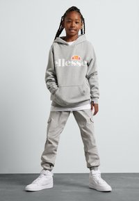 Sudadera gris con logo blanco, pantalones cargo grises y zapatillas blancas de caña alta. Peinado trenzado, de pie contra un fondo neutral.