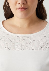 Bílá blůza s texturovaným designem krajky ve vrchní části, hladká dolní část a kulatý výstřih.