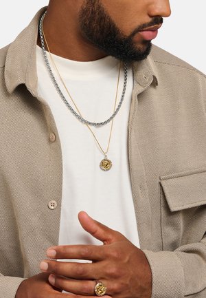 Man wearing beige button-up shirt over white tee, two necklaces—one silver chain, one gold with round pendant—and matching gold ring with round design.