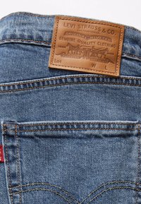 Vaqueros de mezclilla azul con un parche de cuero marrón que presenta texto en relieve y un diseño de caballos. Incluye costuras naranjas y etiqueta de logo.
