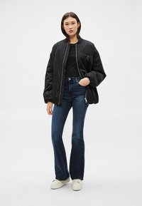 Jauna sieviete, stāvot, ģērbta melnā bombera jakā, melnā topā, tumši zilās zvana biksēs un baltos sporta apavos uz vienkārša fona.