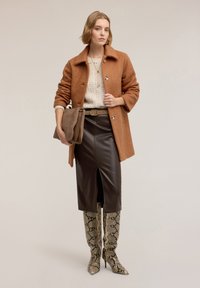 Brązowy płaszcz o teksturowanej powierzchni, kremowy sweter z warkoczowym splotem, dopasowana brązowa skórzana spódnica oraz kozaki za kolano w wężowy wzór z szpiczastymi czubkami. Trzyma brązową torbę.