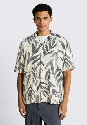 Junger Mann, der ein beige kurzärmeliges Hemd mit dunkelgrauen Blattmustern und dunkle Hosen trägt, steht vor einem schlichten Hintergrund.