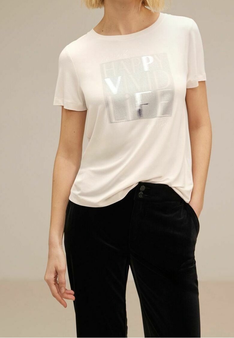 Femme portant une chemise blanche à manches courtes avec un texte argenté brillant "HAPPY VND LIFE" et un pantalon noir, une main dans la poche, sur un fond neutre.