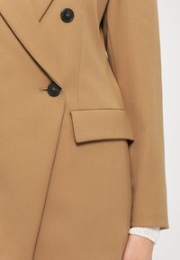 Brun skreddersydd blazer med dobbeltradet design, svarte knapper og skrå lommer. Myk stoff med en glatt tekstur.