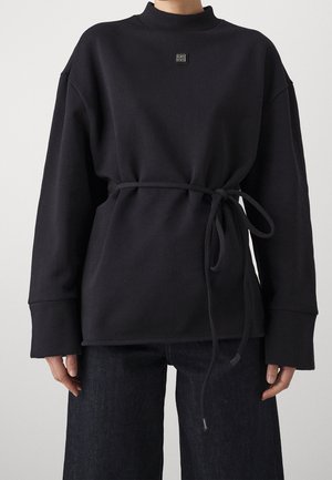 Person trägt einen schwarzen Langarmpullover mit einem Gürtel, der in der Taille gebunden ist, und schwarze Hose vor einfachem hellem Hintergrund.