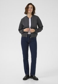 Graue Bomberjacke mit rundem Halsausschnitt, Reißverschluss vorne und gerippten Bündchen. Kombiniert mit dunkelblauer Jeans und schwarzen Schuhen, was einen lässigen Look ergibt.