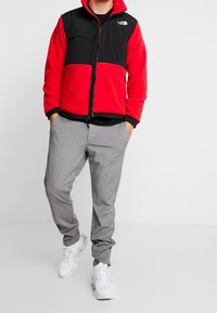 Giacca in pile rossa e nera con chiusura a zip, due tasche sul petto; pantaloni atletici grigi; sneaker bianche; modello in piedi con le mani in tasca.