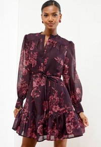 Donna che indossa un abito floreale trasparente a maniche lunghe di un tono di viola scuro, con vita stretta e orlo arricciato, in posa contro uno sfondo bianco semplice.