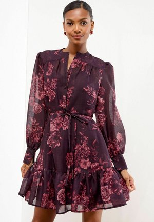 Femme portant une robe florale mauve foncé à manches longues transparentes, avec une taille cintrée et un ourlet à volants, posant devant un fond blanc uni.