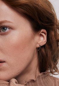 Mały, czarny kolczyk w kształcie kółka noszony w płatku ucha. Zbliżenie na skórę z widoczną teksturą, jasnymi piegami i żeberkowym, beżowym kołnierzykiem swetra.