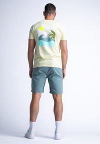 Geel katoenen t-shirt met een grafische print van golven en zon in blauw en groen, gecombineerd met teal shorts en witte sneakers.