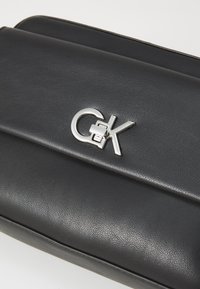Borsa a tracolla in pelle nera con una texture liscia, caratterizzata da una chiusura con logo argentato e un design elegante e strutturato con un pattina piegata.
