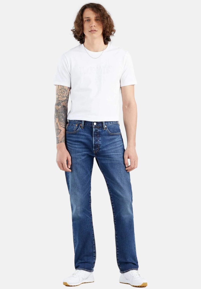 Camiseta de algodón blanca con un logo sutil, combinada con jeans de mezclilla azul. Los jeans tienen un corte recto clásico y un diseño de cinco bolsillos.