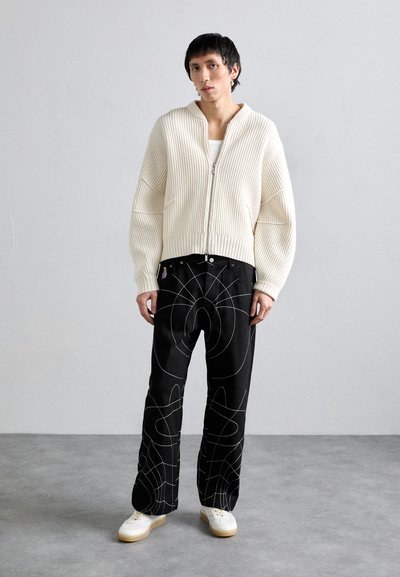 Pulover crem cu dungi, pantaloni negri cu modele abstracte albe și încălțăminte albă cu talpă bej, prezentate pe un fundal neutru.