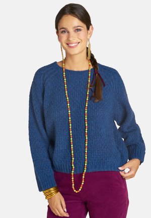 Frau mit geflochtenem Haar, die einen blauen strukturierten Pullover, lange mehrfarbige Perlenkette, goldene Ohrringe, goldene Armbänder und lila Hosen trägt und lächelt.