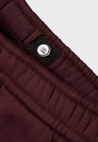 Burgundy byxor med en texturerad midja, prydda med en silverknapp och subtila sömnadsdetaljer, sedda från en vinklad vinkel.