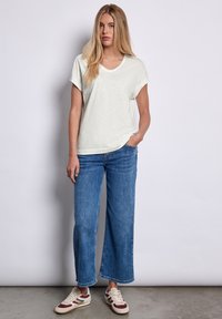 Lys grå V-hals t-shirt med rullede ærmer, parret med brede ben blå denim jeans. Modellen har beige sneakers på med røde og marineblå akcenter.