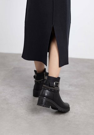 Botines negros de cuero, con un tacón grueso, detalles de cadena y acentos de hebilla, combinados con una falda de punto negra.
