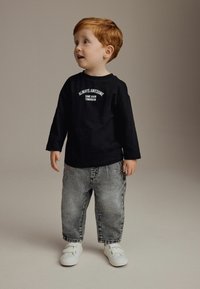 Chemise noire à manches longues avec du texte blanc, associée à un jean gris ample et des baskets blanches. Un garçon se tient de côté, regardant vers le haut.