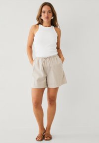 Witte tanktop met ribbeltextuur, gedragen met lichtbeige, loszittende shorts met een elastische tailleband en zijzakken. Bruine sandalen.