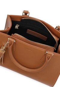 Brūna ādas rokassoma ar dubultiem rokturiem, zelta rāvējslēdzējiem un pušķa piekariņu, parādot plašu melnu iekšpusi un iekšējo rāvējslēdzēja kabatu.